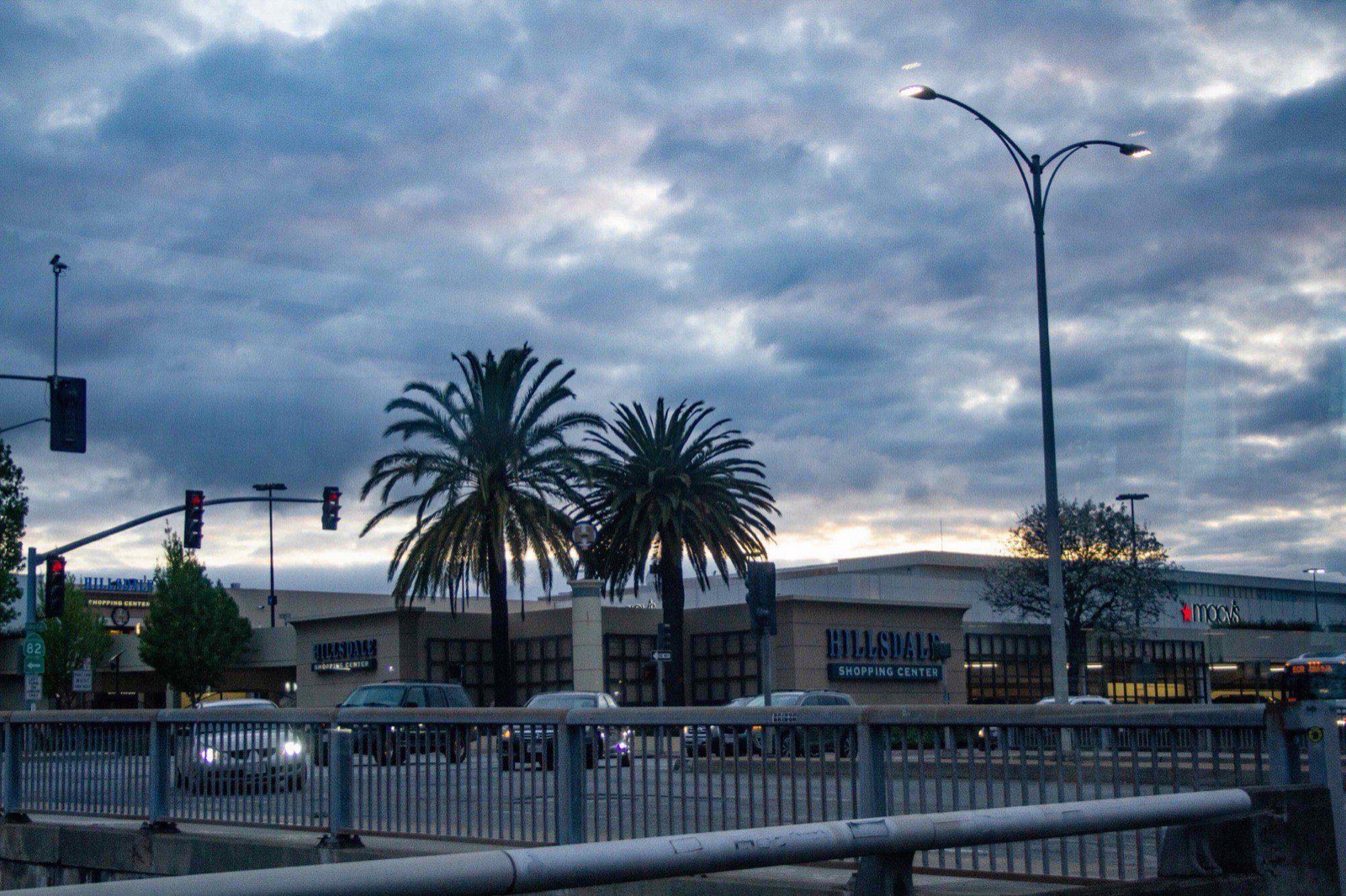 Hillsdale Shopping Center under heavy overcast at dusk, seen across the road from behind a pedestrian railing, with palms and a Macy's sign