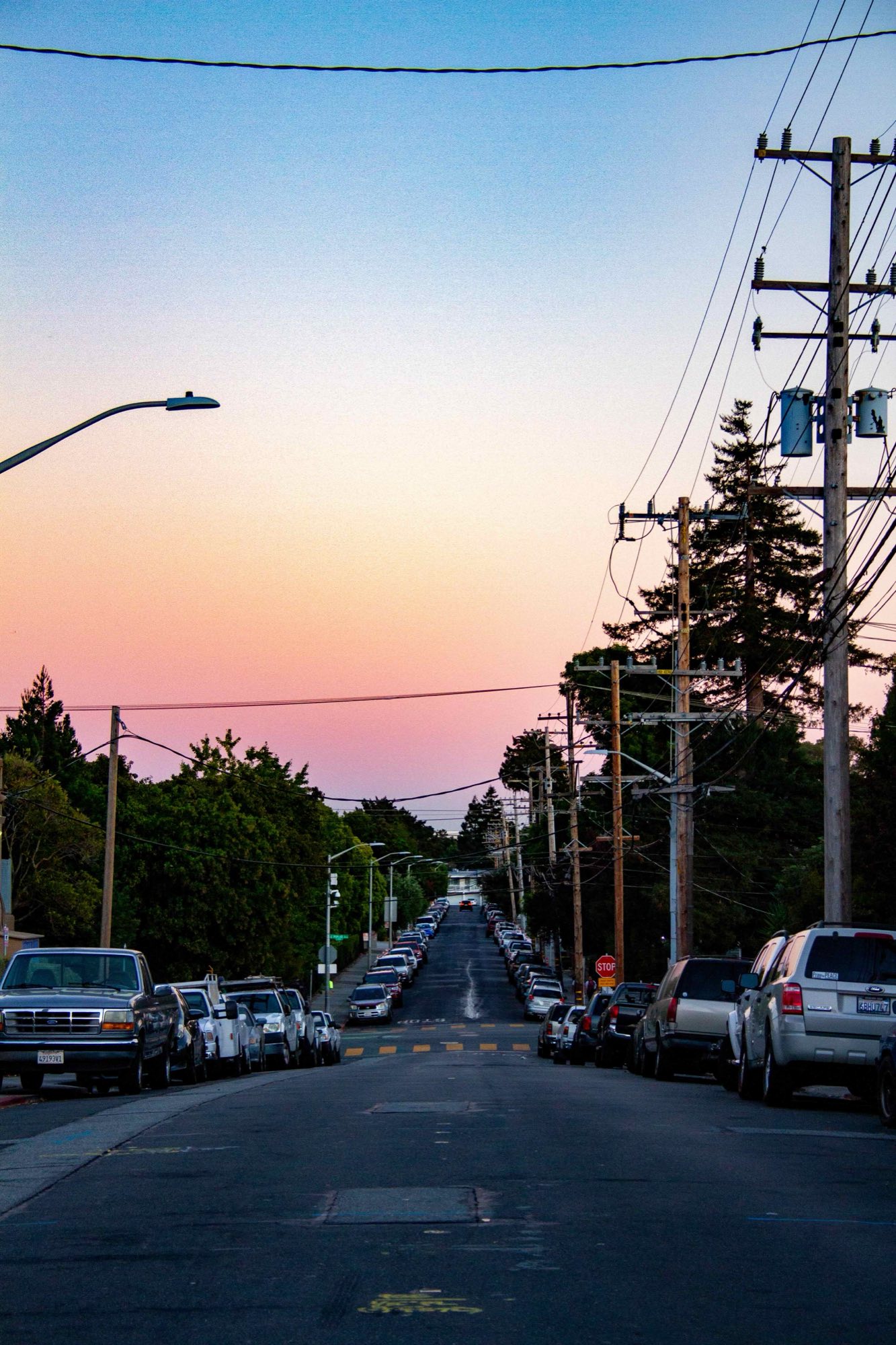 A North Central street at dusk with parked cars filling both curbs the whole way down the block, under a heavy run of utility poles and overhead wires, the sky going orange behind
