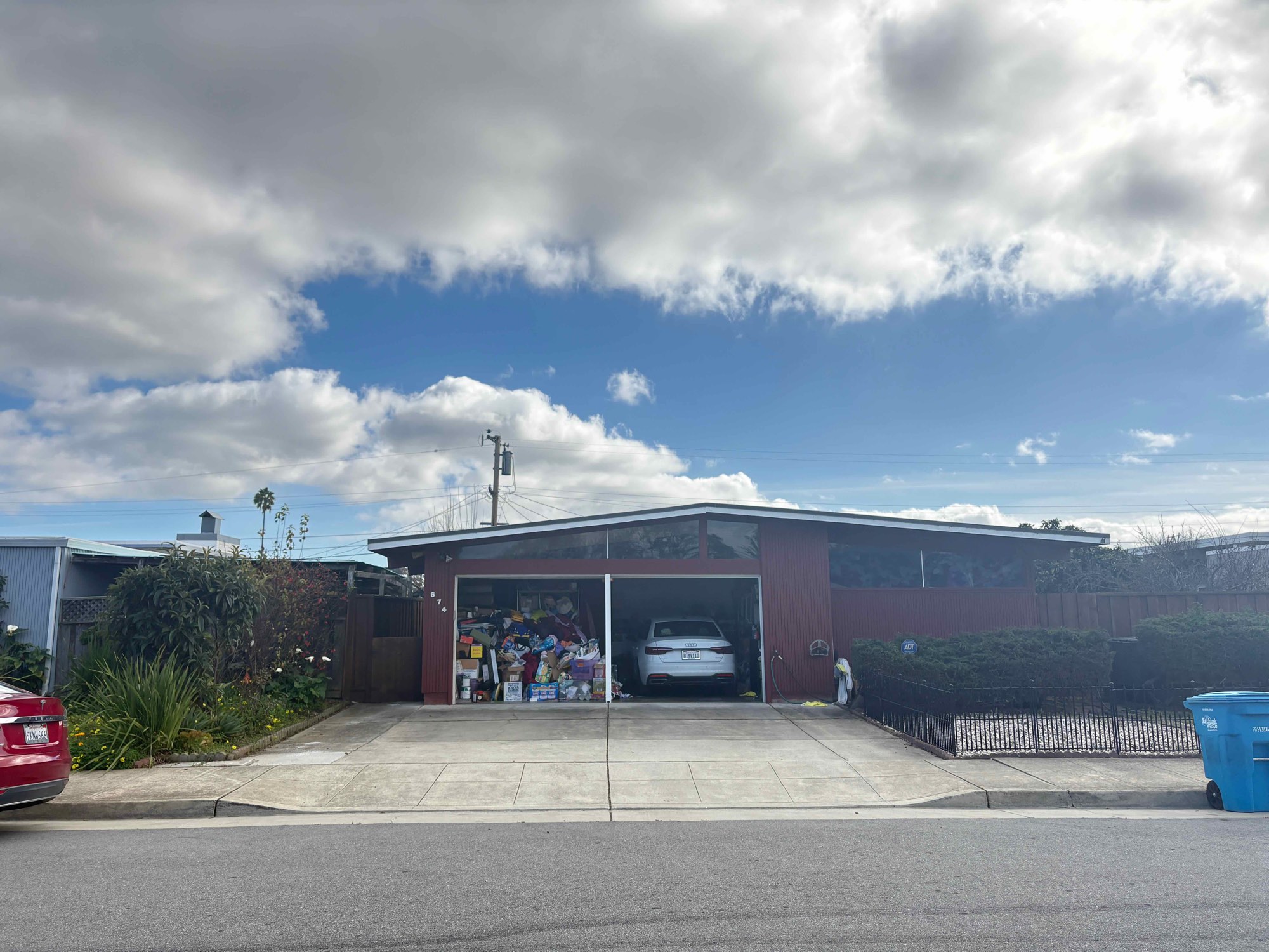 A low mid-century house in Sunnybrae under a broken sky — shallow-pitched roof, a band of clerestory windows above a red board-and-batten front, and a wide concrete driveway running to a double garage