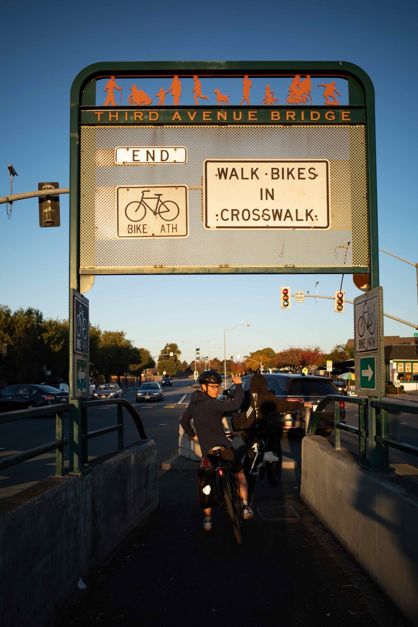 The Third Avenue bridge over US-101 at low sun, seen from the walkway beneath its signs, with a cyclist and a person pushing a stroller crossing toward the far side